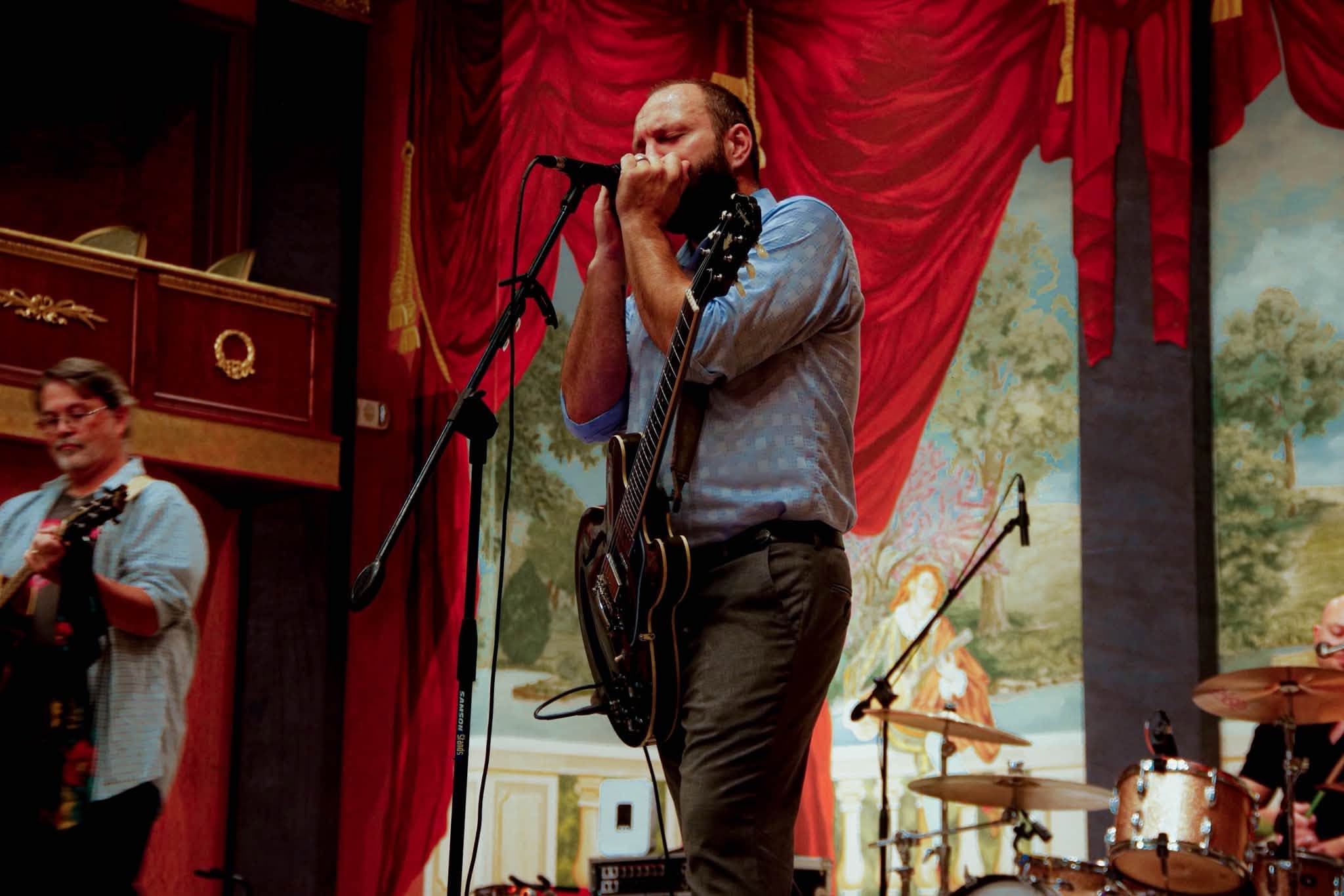 Nate Kelley singing while playing acoustic guitar and harmonica onstage